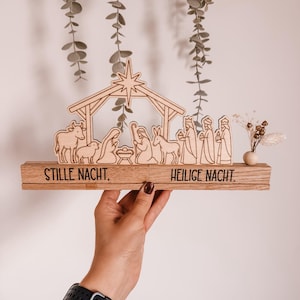 Könnte beinhalten: Eine Krippenszene aus hellem Holz mit einem Stall, einem Stern sowie Figuren von Maria, Josef, dem Jesuskind, Tieren und den Heiligen Drei Königen. Der Sockel besteht aus zwei Holzblöcken mit dem schwarzen Text STILLE NACHT und HEILIGE NACHT.