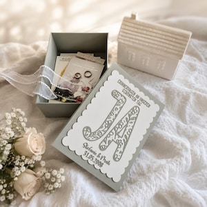 May include: A gray keepsake box with a white scalloped lid featuring the German text JA, ERINNERUNGEN AN UNSEREN SCHÖNSTEN TAG, Stefanie & Maik, and 31.05.2008. Inside are two silver rings on a card, accompanied by a white ceramic house and roses.