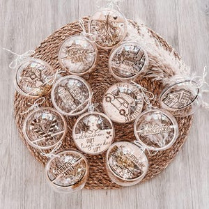 Può includere: Una collezione di palline di Natale trasparenti con vari disegni e testi tagliati al laser, tra cui un omino di pan di zenzero, un angelo e un'auto. Gli ornamenti sono riempiti di glitter dorati e sono disposti su un tappetino intrecciato.