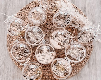 May include: A collection of clear Christmas ornaments with various laser-cut designs and text, including a gingerbread man, angel, and car. The ornaments are filled with gold glitter and are arranged on a woven mat.