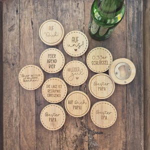 May include: A collection of wooden coasters with engraved text and a bottle opener. The coasters have various phrases, including "Auf uns!" and "Bester Papa." A green glass bottle rests on the wooden surface.