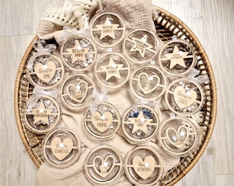 May include: A wicker basket filled with clear, round ornaments with wooden cutouts and text. The ornaments have various designs, including stars and hearts, and phrases like "Best Dad" and "Best Grandma". The basket is lined with a beige cloth.