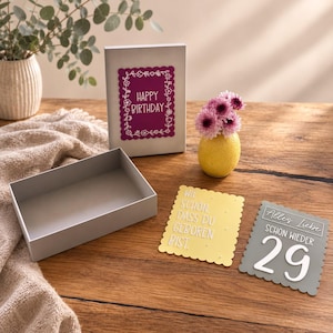 May include: A light gray gift box with a lid featuring a magenta scalloped card that says Happy Birthday. Beside it are a yellow scalloped card with the text Wie schön, dass du geboren bist and a gray card with the text Alles Liebe schon wieder 29.