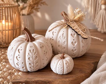 May include: Three white decorative pumpkins with brown stems, featuring embossed heart and leaf patterns. The largest pumpkin has a small wooden tag tied to its stem with twine, displaying the German text Herbstliebe and dried floral accents.