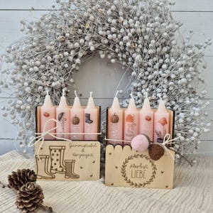 May include: Two sets of decorative candles in wooden boxes, featuring fall-themed illustrations and text. The candles are pink and adorned with images of boots, pumpkins, and mushrooms. A white wreath and pinecones are in the background.