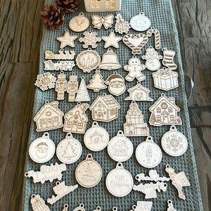 Puede incluir: Una colección de adornos navideños de madera en varias formas, incluyendo estrellas, copos de nieve, casas y árboles de Navidad. Algunos adornos tienen diseños grabados y texto. También hay un portavelas blanco en forma de casa.