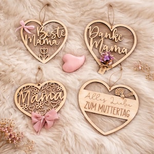 Op de afbeelding: Vier houten hartvormige ornamenten met tekst en decoratieve elementen. Eén zegt "Beste Mama" met een roze strik, een andere zegt "Danke Mama" met bloemen, een derde zegt "Mama" met een roze strik, en de laatste zegt "Alles Liebe ZUM MUTTERTAG".