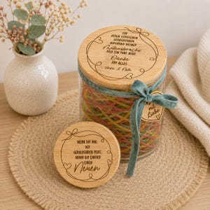 May include: A clear glass jar filled with colorful yarn, topped with a round wooden lid engraved with German text and a decorative border. A second matching wooden lid sits in front, also engraved with German text. A teal ribbon is tied around the jar.