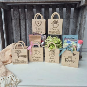 Könnte beinhalten: Sechs helle Geschenktaschen aus Holz mit Griffen, jede mit individuellen Lasergravuren und deutschem Text. Die Taschen zeigen Motive wie ein Herz, eine Gießkanne, einen Blumenstrauß, eine Kaffeetasse, ein Glas und ein Herz mit dem Text Für Dich.
