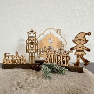 Könnte beinhalten: Weihnachtsdekorationen aus Holz mit einem Nussknacker, einem Elf und einem Lebkuchenhaus. Die Wörter "Frohe Weihnachten" und "Winter Zauber" sind ebenfalls abgebildet. Die Dekorationen stehen auf einer braunen Basis mit Tannenzapfen und Grün.