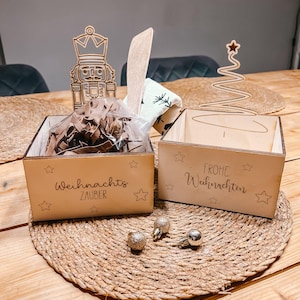 May include: Two wooden boxes with laser-cut text and star designs. One box reads "Weihnachts ZAUBER" and the other says "FROHE Weihnachten". The boxes sit on a woven mat with gold and silver ornaments. A wooden Christmas tree and nutcracker are in the background.