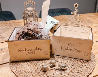 May include: Two wooden boxes with laser-cut text and star designs. One box reads "Weihnachts ZAUBER" and the other says "FROHE Weihnachten". The boxes sit on a woven mat with gold and silver ornaments. A wooden Christmas tree and nutcracker are in the background.