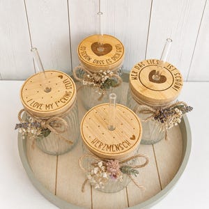 Op de afbeelding: Vier glazen potten met houten deksels en doorzichtige rietjes, gerangschikt op een rond dienblad. De deksels hebben gegraveerde tekst en hartontwerpen. Elke pot is versierd met touw en gedroogde bloemen. De tekst bevat "I LOVE MY F*CKING SELFI", "SO SCHON, DASS ES DICH GIBT!", "WER DAS LIES IST SCHON!" en "HERZMENSCH".