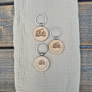 Puede incluir: Tres llaveros de madera con anillos plateados. Cada llavero presenta un colgante circular de madera con un diseño de camión de bomberos y la palabra "Feuerwehrmann" grabada alrededor del borde. Los llaveros están sobre una tela beige.