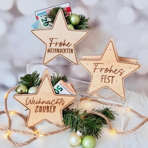May include: Three light-coloured wooden star-shaped holders with engraved text. The stars feature the phrases Frohe WEIHNACHTEN, Frohes FEST, and Weihnachts ZAUBER. Each holder contains green pine branches, small ornaments, and paper currency.