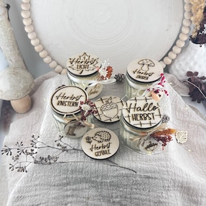Könnte beinhalten: Vier Gläser mit Holzdeckeln, die mit herbstlichen Motiven und deutschen Schriftzügen wie Herbst Licht, Herbst Zauber, Herbst Knistern und Hallo Herbst graviert sind. Jeder Deckel ist mit kleinen Trockenblumen und Bindfaden verziert.