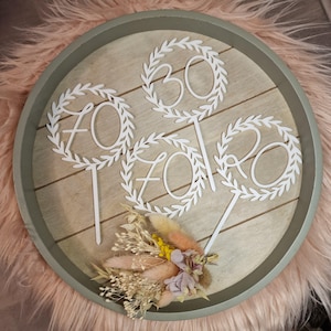 Könnte beinhalten: Ein Kuchenaufsatz aus Holz mit den weißen Zahlen 70, 30 und 20, jeweils umgeben von einem Lorbeerkranz-Design. Der Aufsatz befindet sich auf einer runden Holzbasis mit einem Trockenblumenarrangement und einem grünen Rand.