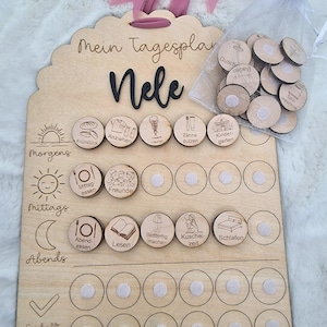 Peut inclure: Un tableau de planification quotidienne en bois avec le nom "Nele" en écriture noire. Le tableau comporte des icônes gravées pour les activités quotidiennes et est livré avec des jetons en bois dans un sac en filet. Un ruban rose est attaché en haut.