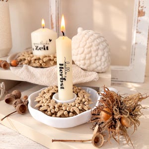 Könnte beinhalten: Eine cremefarbene Stabkerze mit dem schwarzen Aufdruck "hallo Herbst", die in einem weißen Kerzenhalter aus Keramik steht, welcher mit einem Kranz aus kleinen beigen Beeren und Blättern verziert ist, mit einer brennenden Flamme.