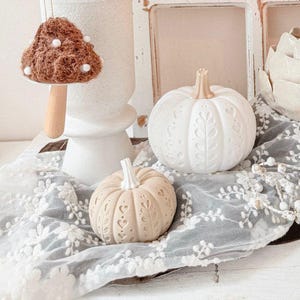 May include: A decorative display featuring two carved pumpkins with intricate leaf and heart patterns, a hanging brown textured mushroom ornament with white dots, a white ceramic vase, and a white floral lace fabric draped underneath.