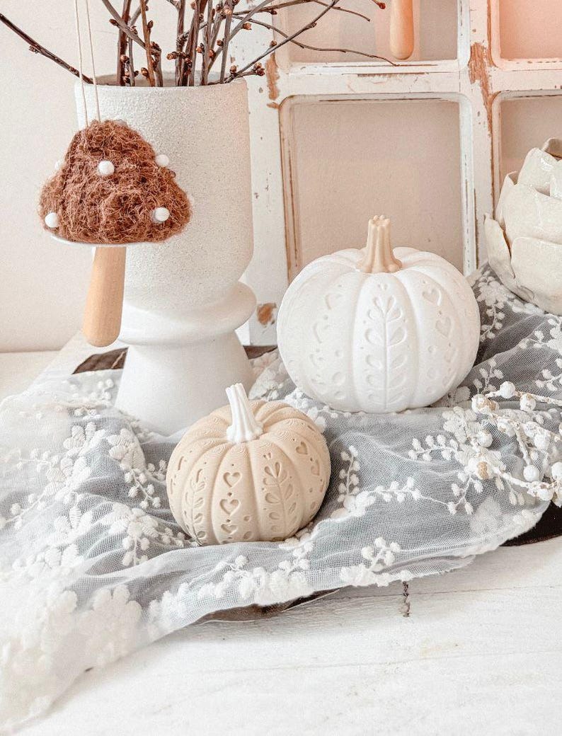 May include: A decorative display featuring two carved pumpkins with intricate leaf and heart patterns, a hanging brown textured mushroom ornament with white dots, a white ceramic vase, and a white floral lace fabric draped underneath.