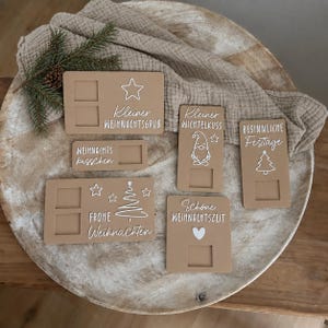 May include: Five tan rectangular plaques with white text and icons including stars, a gnome, a heart, and Christmas trees. The plaques feature cut-out squares and phrases such as Kleiner Weihnachtsgrub, Weihnachtskusschen, Frohe Weihnachten, Kleiner Wichtelkuss, Besinnliche Festtage, and Schone Weihnachtszeit.