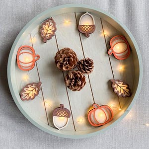 May include: A round, light green wooden tray containing three brown pinecones and a string of LED fairy lights. The lights are woven through wooden cutouts shaped like pumpkins, acorns, and leaves, which glow with a warm, soft illumination.