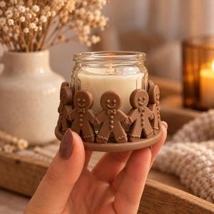 Op de afbeelding: Een kleine glazen kaarsenhouder met een ronde voet versierd met een ring van bruine peperkoekmannetjes. De doorzichtige glazen pot bevat een witte waskaars met een zichtbare lont, wat zorgt voor een decoratief accessoire voor op tafel.