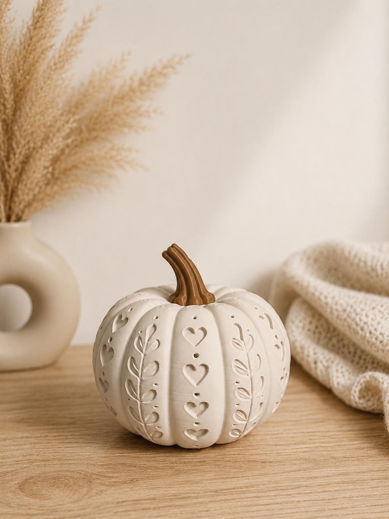 May include: A cream-coloured decorative pumpkin featuring a brown stem and intricate cut-out patterns. The surface is adorned with alternating vertical sections of heart shapes and leaf-like vine designs, creating a hollow, textured appearance.