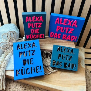May include: Four rectangular signs in shades of blue with pink or black lettering. Two signs read "ALEXA, PUTZ DIE KÜCHE!" and two signs read "ALEXA, PUTZ DAS BAD!" The signs feature either raised pink lettering or black cut-out text on blue backgrounds.