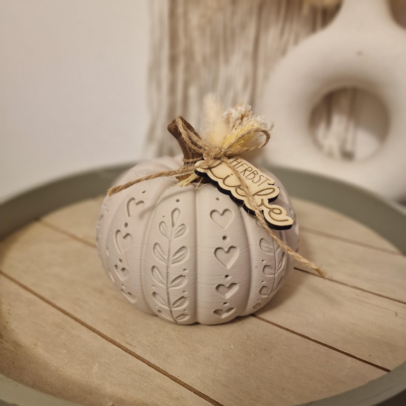 May include: A light grey decorative pumpkin featuring embossed heart and leaf patterns. The pumpkin has a brown stem wrapped in twine, a small bunch of dried beige grass, and a wooden tag with the text Herbstliebe attached to the stem.