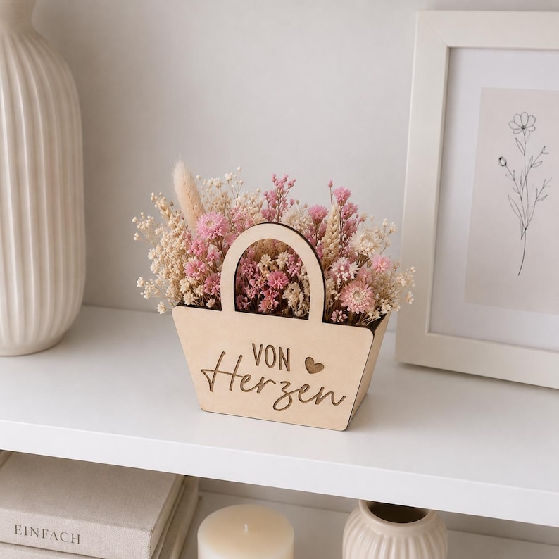 May include: A light-colored wooden basket with a handle, featuring the engraved text "VON Herzen" and a small heart symbol. The basket is filled with a dense arrangement of dried flowers in shades of pink, cream, and beige, including baby's breath.
