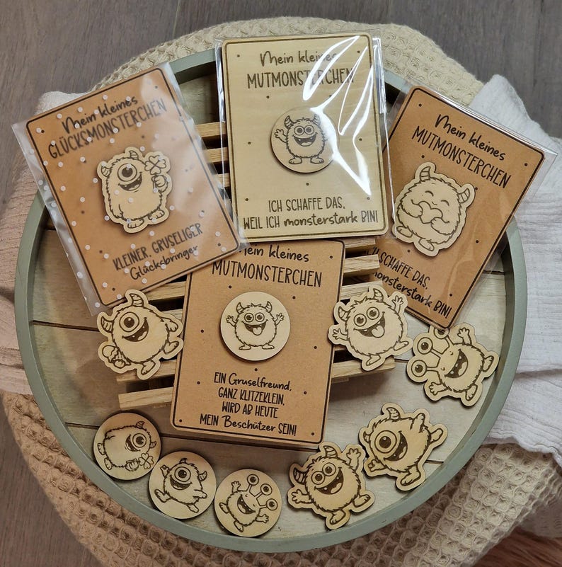 May include: A collection of wooden monster-themed pins and cards. The pins feature various cartoon monster designs. The cards have text in German, including "Mein kleines MUTMONSTERCHEN" and "KLEINER, GRUSELIGER Gl&uuml;cksbringer."