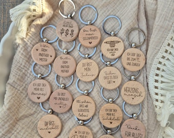 May include: A collection of wooden keychains with silver key rings. Each keychain has a circular wooden pendant with engraved text and small designs. The phrases include "Sister from another mister" and "I love you more than pizza."