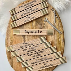 Puede incluir: Reglas plegables de madera con detalles metálicos dorados y texto negro dispuestas sobre una tabla ovalada de madera. Las reglas incluyen frases como "Superpapa!" y "Papa, Kann Alles Reparieren!" Una llave inglesa y clavos también están presentes.