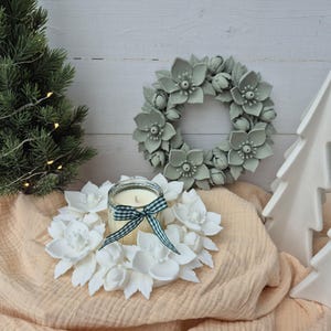 Puede incluir: Un frasco de vela de cristal con cera color crema y una cinta de cuadros verdes y blancos, colocado dentro de una corona floral blanca. Una corona floral verde salvia cuelga en la pared, junto a un pequeño pino artificial y árboles blancos.
