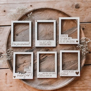 May include: Six rectangular light-colored wooden photo frames with various German phrases and small heart icons engraved on the bottom borders. The frames feature a clean, simple design and are arranged on a round wooden tray with dried flowers.