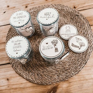 Puede incluir: Un conjunto de tarros de cristal con tapas de madera y posavasos decorativos. Los tarros tienen texto en alemán, incluyendo "DU BIST MIR UNENDLICH wichtig" y "HERZ mensch". Los posavasos también tienen texto e ilustraciones en alemán.