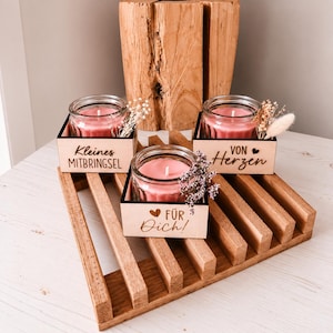 Könnte beinhalten: Drei Kerzen in Glasbehältern mit rosa Wachs, jeweils in einer quadratischen Holzbox. Die Boxen tragen die Aufschriften "Kleines MITBRINGSEL", "VON Herzen" und "FÜR Dich!" mit kleinen Herzsymbolen. Trockenblumen sind in jedem Glas arrangiert.