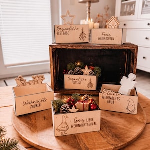 Könnte beinhalten: Eine Sammlung von hellen Holzkisten mit verschiedenen weihnachtlichen, lasergravierten Texten und Illustrationen, darunter eine Gans, ein Wichtel, ein Schlitten und Tannenzapfen, teilweise gefüllt mit kleinen Tannenzweigen und Dekorationen.