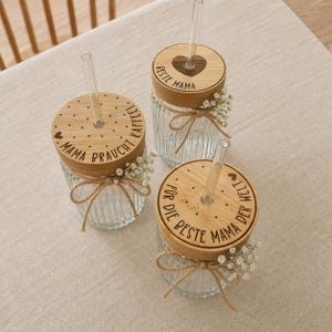 Op de afbeelding: Drie glazen drinkglazen met bamboe deksels en glazen rietjes. De deksels hebben tekst en hartmotieven. Elke pot is versierd met een touwstrik en kleine witte bloemen. De tekst op de deksels luidt "Mama braucht Kaffee" en "Für die beste Mama der Welt."