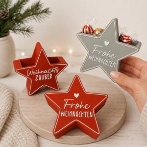 Könnte beinhalten: Drei sternförmige Behälter mit offener Oberseite. Zwei sind rot mit weißem Text "Weihnachts ZAUBER" und "Frohe WEIHNACHTEN" sowie einem kleinen Herz. Einer ist grau mit "Frohe WEIHNACHTEN" und einem Herz, gefüllt mit eingewickelten Süßigkeiten.