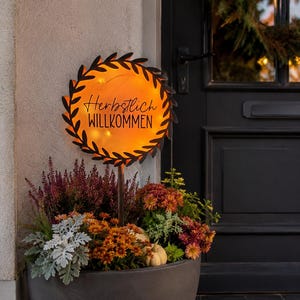 Könnte beinhalten: Ein rundes Metall-Gartenschild mit schwarzem Blattrand und leuchtend orangefarbener Mitte. Das Schild trägt den schwarzen Schriftzug "Herbstlich WILLKOMMEN" und ist auf einem Stab in einem großen grauen Pflanzkübel mit Herbstblumen befestigt.