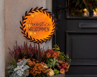 Könnte beinhalten: Ein rundes Metall-Gartenschild mit schwarzem Blattrand und leuchtend orangefarbener Mitte. Das Schild trägt den schwarzen Schriftzug "Herbstlich WILLKOMMEN" und ist auf einem Stab in einem großen grauen Pflanzkübel mit Herbstblumen befestigt.