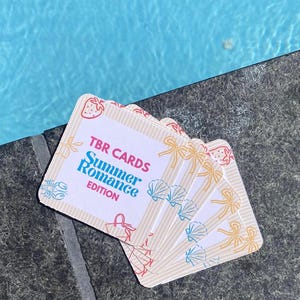 May include: A stack of TBR Cards, Summer Romance Edition, with a light beige striped background. The cards feature illustrations of strawberries, seashells, bows, and a handbag. The text on the cards reads "TBR CARDS Summer Romance EDITION".