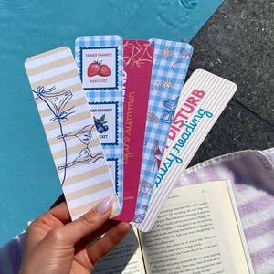 May include: A hand holds a selection of decorative bookmarks. One bookmark reads "DISTURB busy reading". Other bookmarks feature illustrations of strawberries, blueberries, and beach-themed graphics. The bookmarks are of various colours.