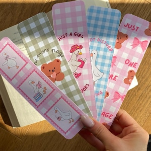 Puede incluir: Una colección de cinco marcapáginas decorativos con diseños caprichosos. Cada marcapáginas presenta un patrón diferente, incluyendo cuadros de vichy e ilustraciones de patos, osos y lazos. El texto en los marcapáginas dice "Just a Girl", "One More Page" y "Not Distrubing".