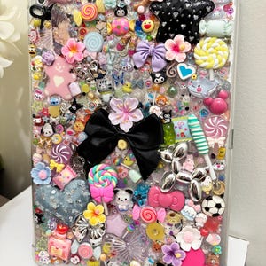 May include: A clear tablet case densely decorated with an assortment of colorful charms. The charms include flowers, bows, candies, stars, and animal figures. The case has a whimsical and playful aesthetic.