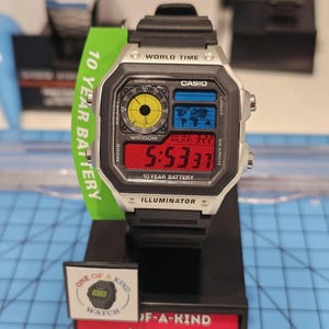May include: A silver and black Casio digital watch with a black band, displaying the time 5:53:37. The watch face includes a world map and the text "10 YEAR BATTERY". A green tag reads "10 YEAR BATTERY".