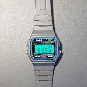 May include: A gray Casio F-91W digital watch with a black strap. The watch face has a blue border and displays the time, day, and date. The watch has the text "CASIO F-91W" and "WATER RESIST".